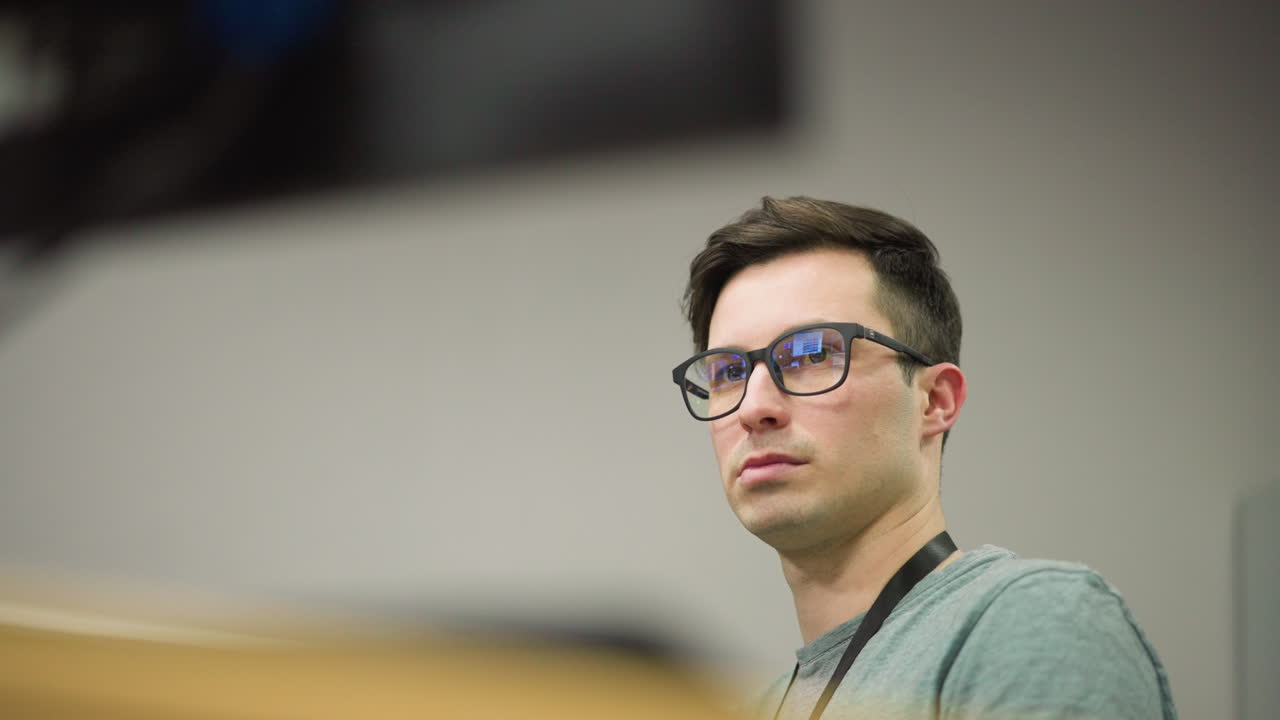 Close-up of young man with glasses sitting in front of computer screen in office environment, looking serious and focused, deep in thought while working on digital task in professional setting
