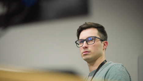 close-up of young man with glasses sitting in front of computer screen in office environment, looking serious and focused, deep in thought while working on digital task in professional setting