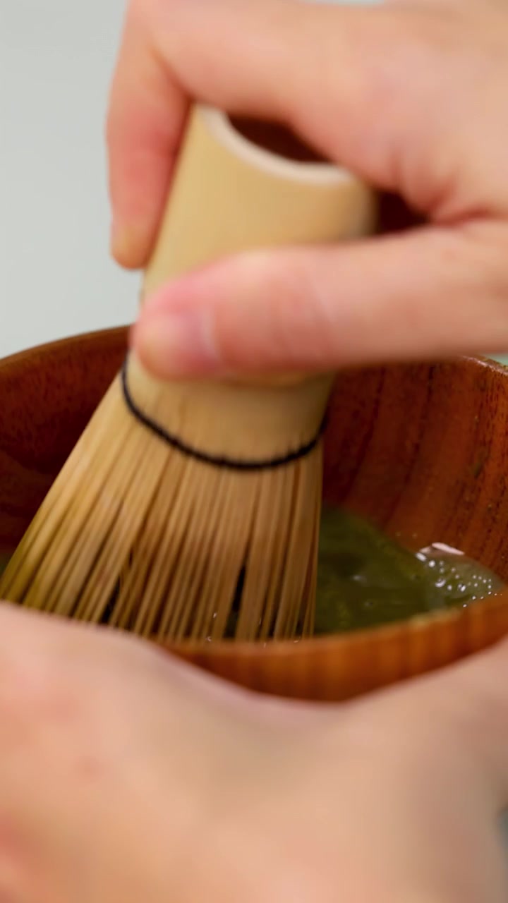 Primo piano di mani che sbattono tè matcha verde in una ciotola di legno con illuminazione da studio soffusa.