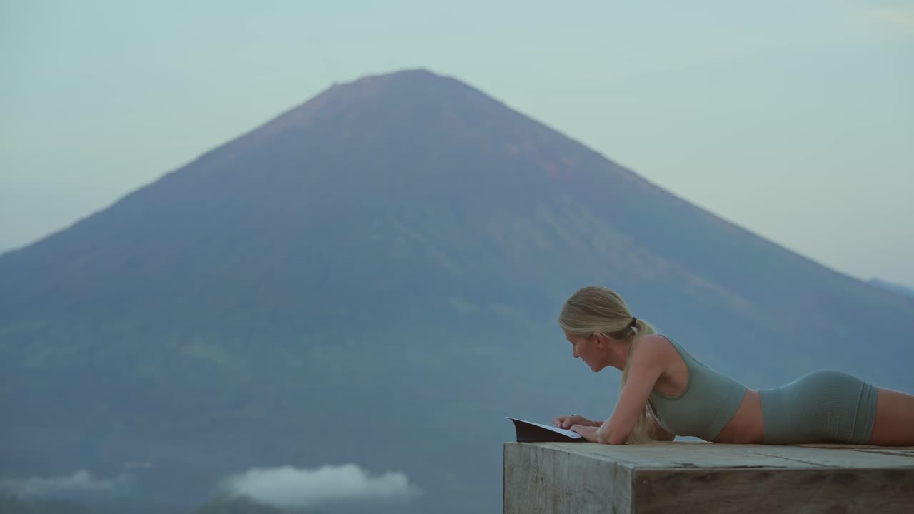Woman laying on platform while writing in journal with mount Agung, self care concept