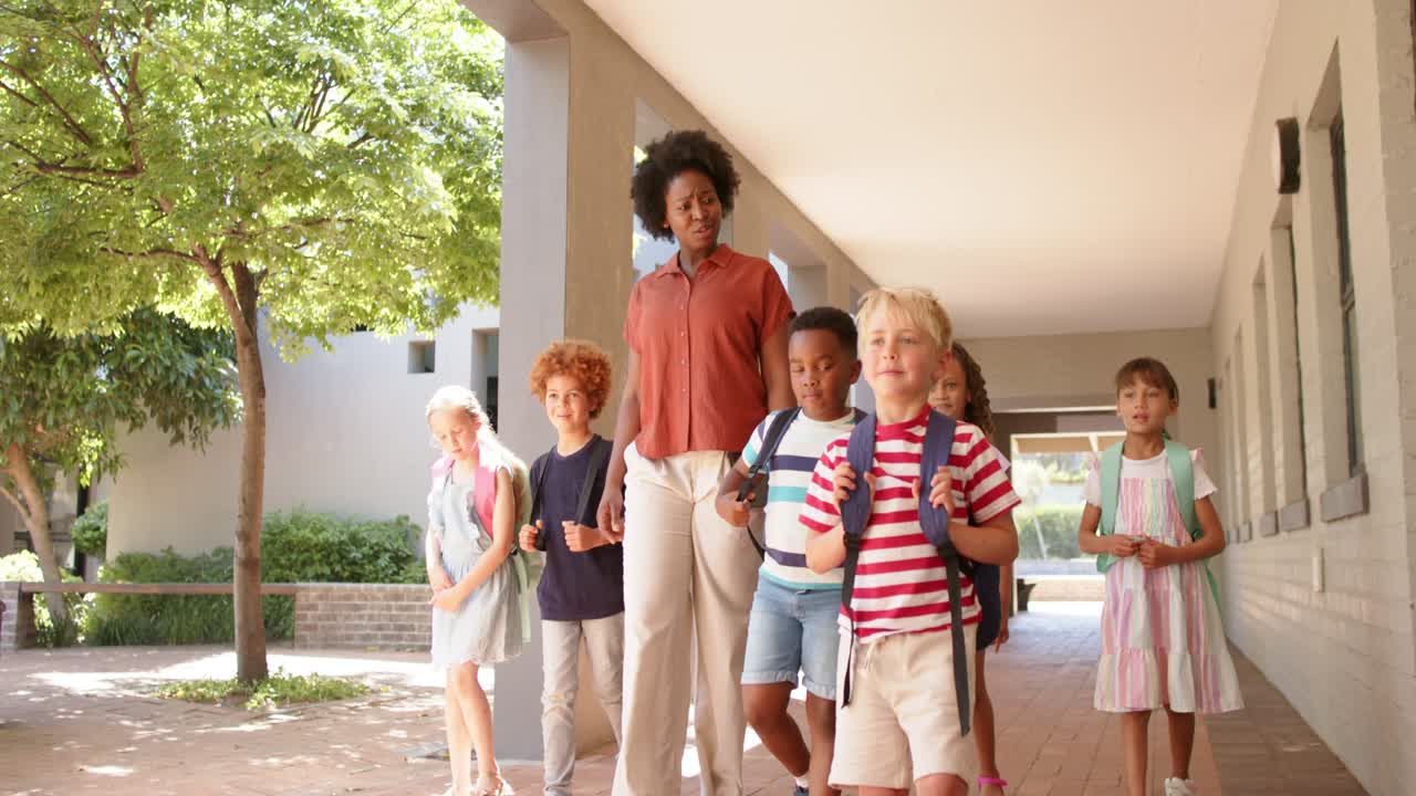 Teacher with children guiding along walkway with columns backpacks as blonde boy stepping to lead