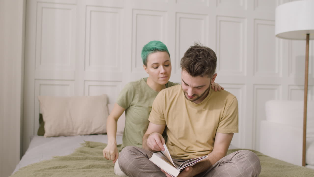 молодой человек сидит на кровати и листает книгу, а его девушка обнимает его