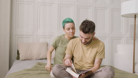 young man sitting on the bed and leafing through a book while his girlfriend cuddling him