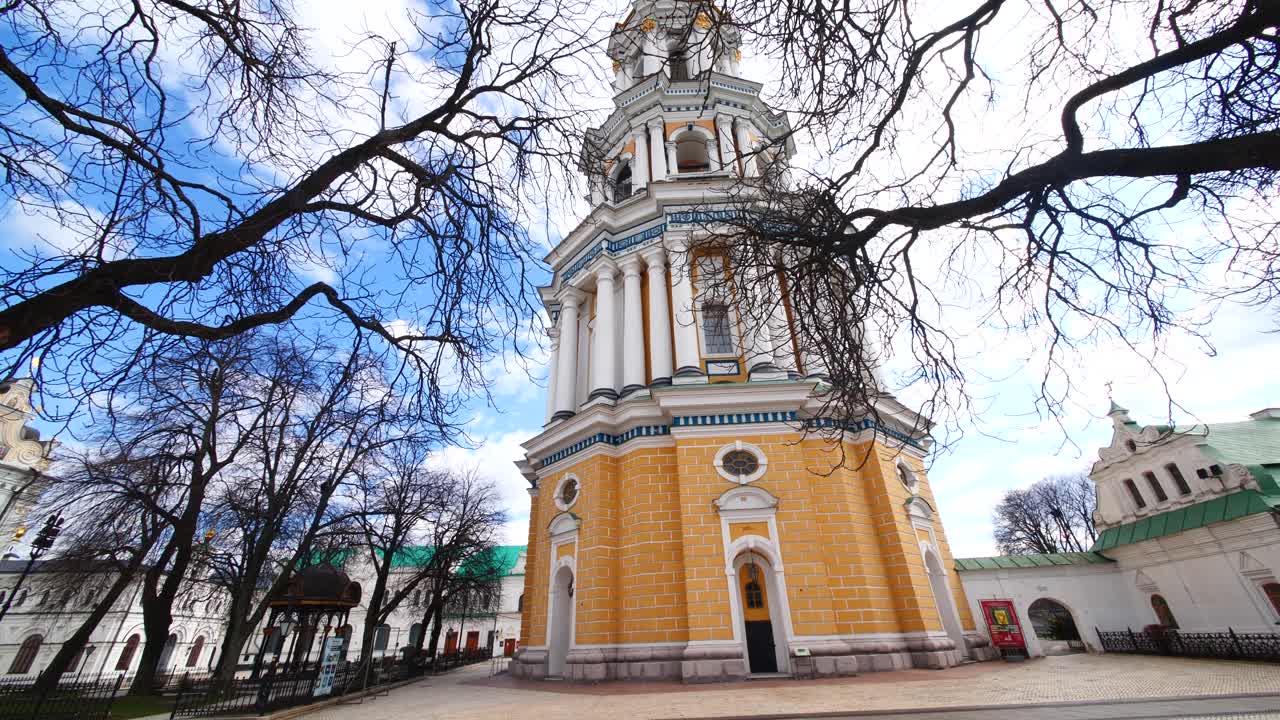 campanario barroco amarillo, azul y blanco detrás de árboles marrones de invierno sin hojas, panorámica hacia abajo, kiev, ucrania, kiev pechersk lavra