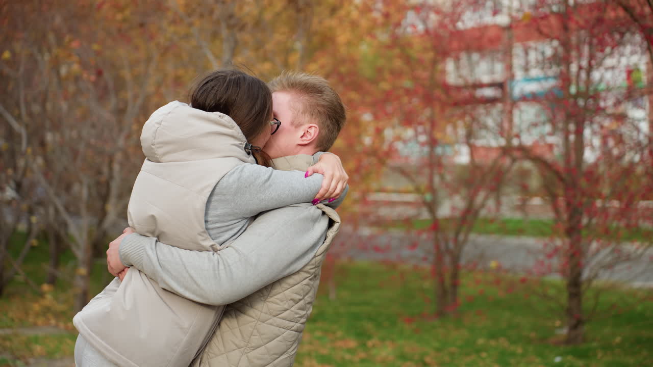 Man and wife hug each other tightly as they share warm kiss outdoors in autumn park, dressed in matching jackets, surrounded by trees with colorful leaves, enjoying romantic moment together