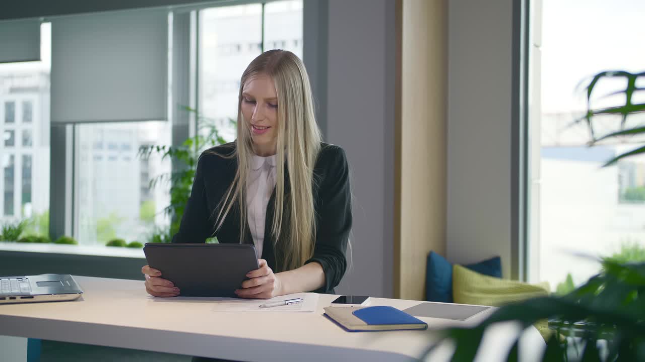 Stilfull forretningskvinne med nettbrett på kontoret. Elegant kvinne i dress sitter ved bordet med bærbar PC og bruker nettbrett på et moderne, lyst kontor med store vinduer.