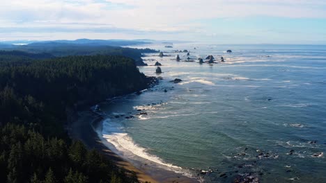 stunning aerial drone dolly out shot of the gorgeous third beach in forks, washington with large rock formations, surrounded by a pine tree forest on cliffs, and golden sand on a warm summer morning.