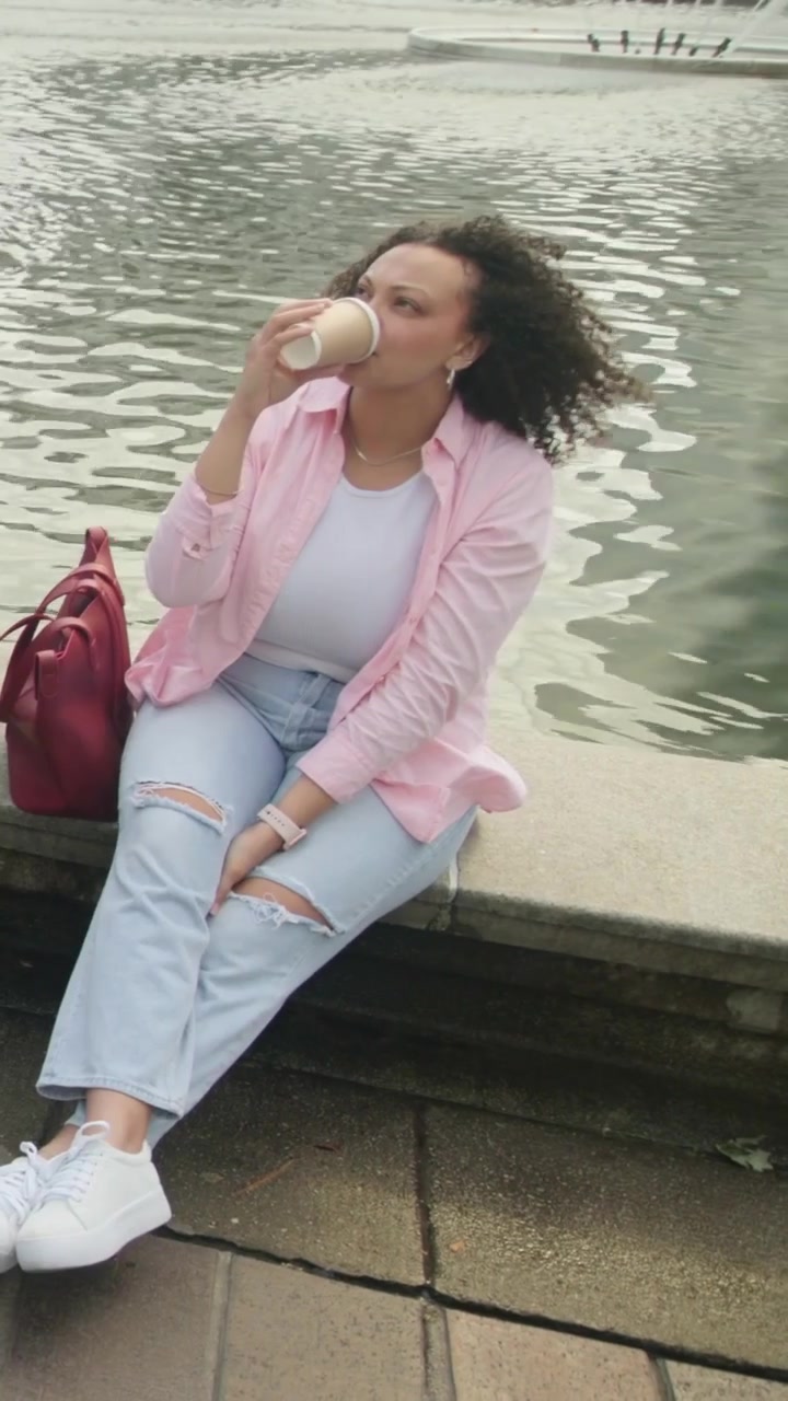 Vídeo vertical: Transeunte passando, mulher de camisa rosa sorrindo, ajeitando o cabelo, bebendo de uma xícara na piscina.