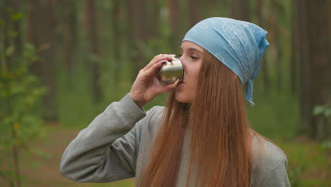 young woman in blue bandana and grey sweater savors drink from thermos cup while relaxing in peaceful forest, partial view of companion in green shirt nearby