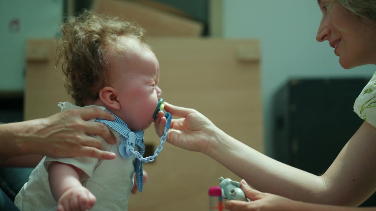 Baby with pacifier held by a parent, looking at colorful toy being offered, representing childhood care and engagement