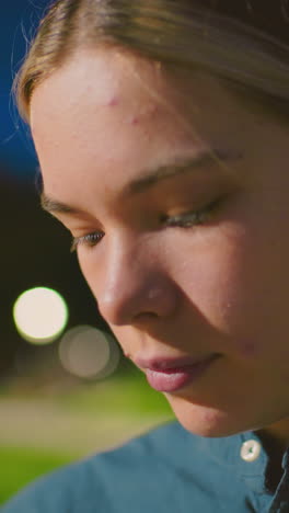 primer plano de una mujer joven en un tranquilo parque al aire libre por la noche, mostrando una expresión pensativa bajo el suave resplandor de las luces, el fondo incluye vegetación borrosa y iluminación suave