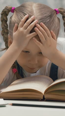 slow motion: cute happy little child with plaits tired of doing home tasks with textbook sits at table in light room at quarantine time
