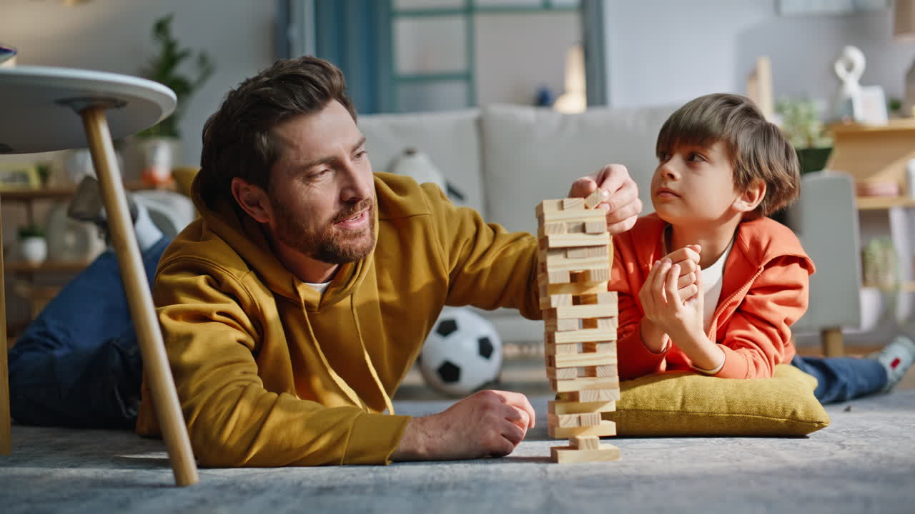 Genitore e figlio che costruiscono una torre e si divertono con Jenga sdraiati insieme sul pavimento dell'appartamento, primo piano.