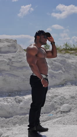 handsome guy without shirt standing on white rocky background. dark skinned guy in black cap and glasses with muscular body outdoors. profile view. vertical video