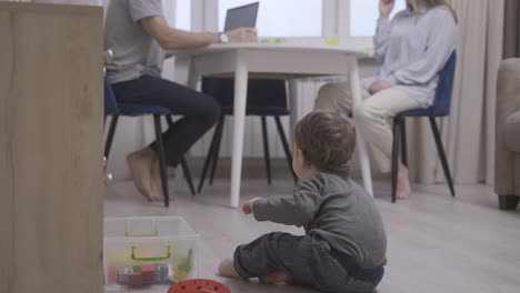 parents sitting at a table while their baby is sitting on the floor playing with toys. then the baby takes his first steps to her mother and she gives him a carrot.