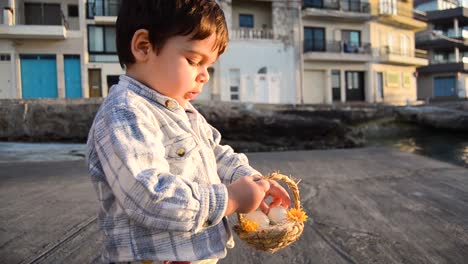 adorable niño pequeño con la iluminación del sol poniente jugando con sus huevos de juguete y su cesta junto al puerto deportivo