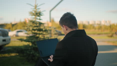 designer seated outdoors operating laptop with leg crossed in serene environment, background includes moving car, nearby parked car, urban structures, and greenery