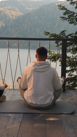 young couple meditates on mat on glamping terrace. focused people do yoga exercises for relaxation together looking at lake. concept of mindfulness lifestyle