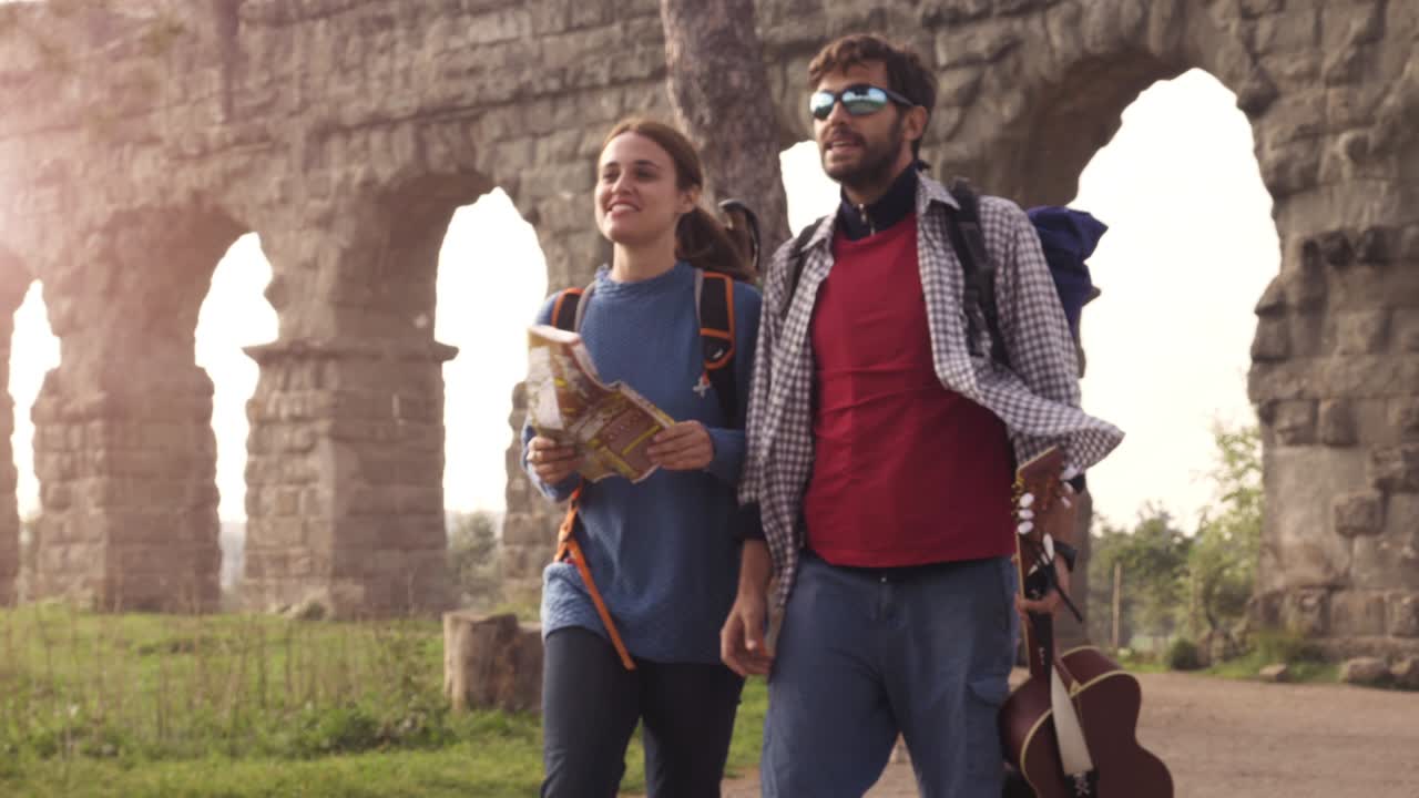 Young couple backpackers tourists walking with map guide near roman aqueduct arches in parco degli acquedotti park ruins in rome at sunrise with guitar and sleeping bag slow motion steadycam