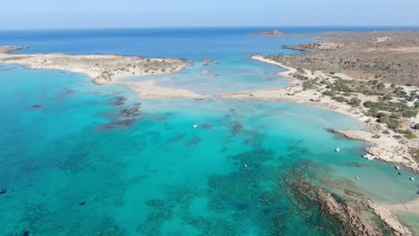 Drone-view-in-Greece-flying-over-Elafonisi-white-sand-narrow-beach,-clear-blue-water-on-the-sides-and-brown-landscape-on-a-sunny-day