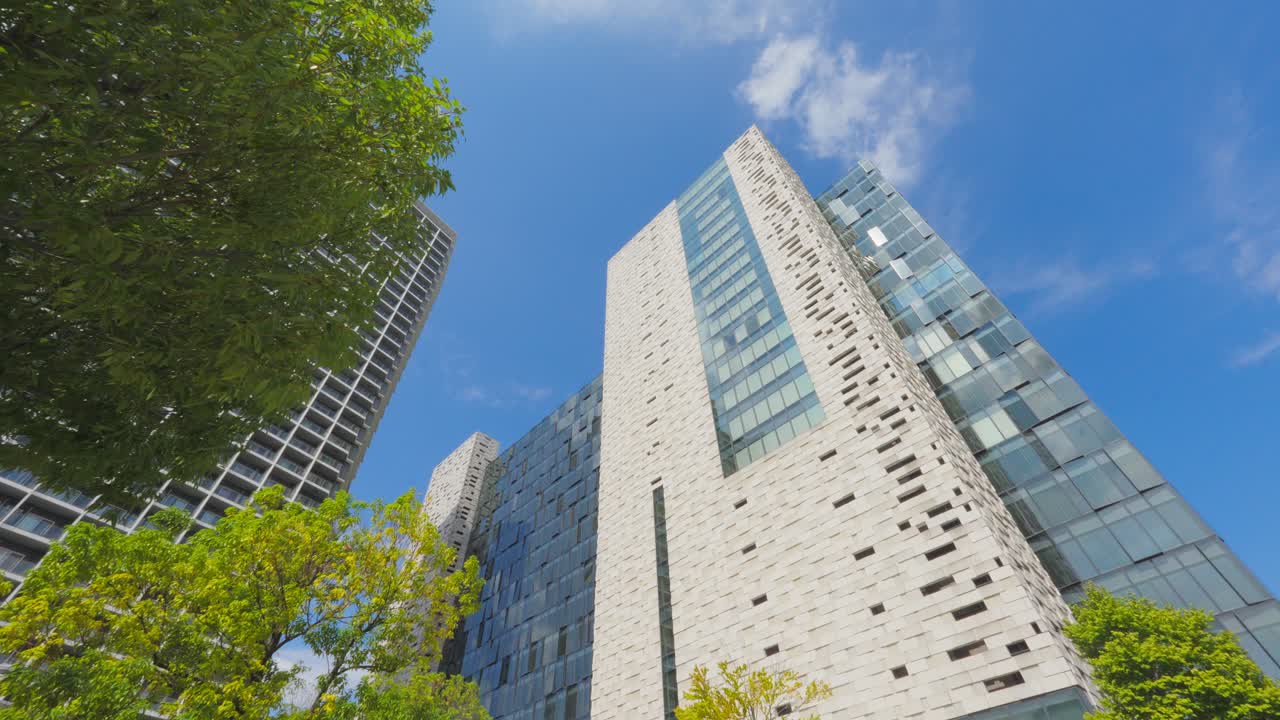 A dramatic low-angle shot of a modern Tokyo skyscraper with contrasting glass and stone facades, framed by lush green trees and a blue sky