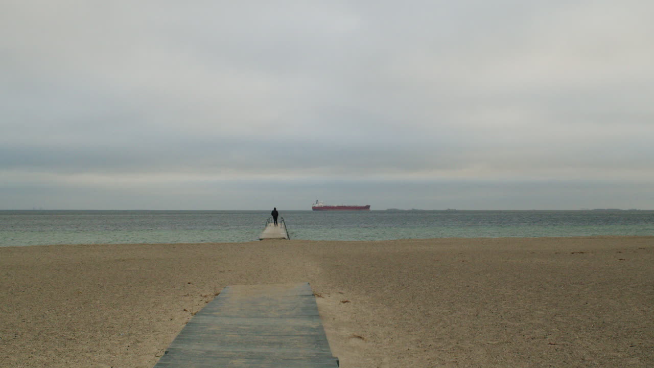 A wide, symmetrical shot of a solitary figure walking away from the camera down a narrow pier or jetty. The path leads the viewer's eye across a vast sandy beach toward the sea