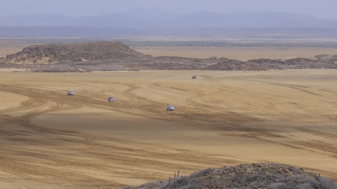 Overview of offroading cars driving along arid Guajira desert in Colombia, vast golden sands stretching under clear skies