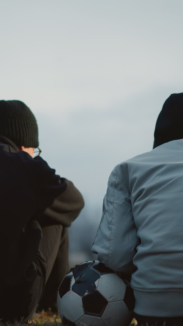 두 소년은 저녁에 잔디에 앉아 있으며, 한 명은 축구공에 왼손을 고 있으며, 빛의 반사와 배경에 빛의 흐릿한 시야와 건물이 있습니다.