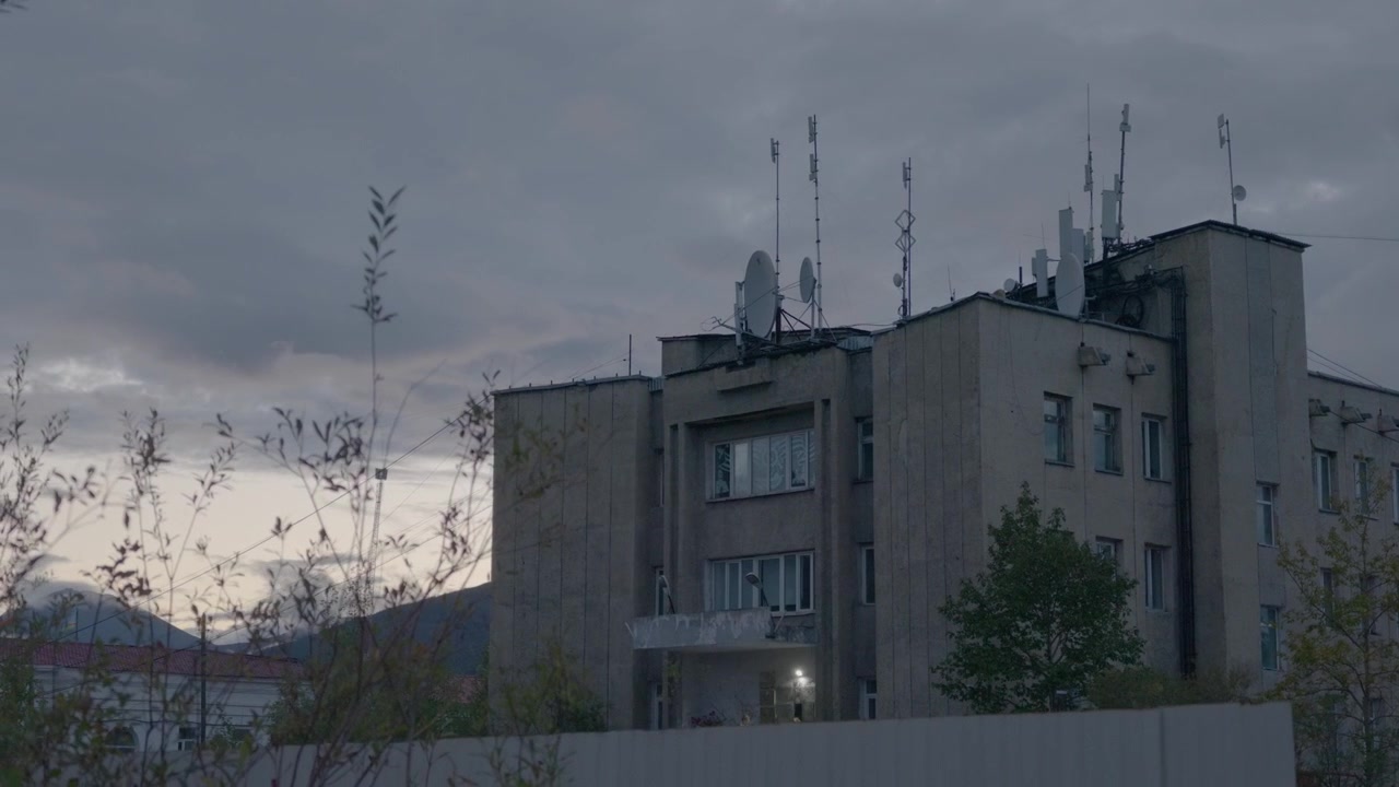 Um edifício de concreto cinza com antenas no telhado é lentamente panoramizado para a esquerda, revelando árvores ao redor e um céu nublado, com uma janela iluminada perto da base.