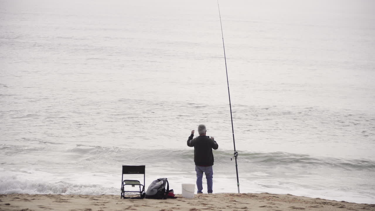 Nelayan Berdiri di Pantai Berpasir dengan Joran Pancing, Memancing di Laut dengan Ombak. - bidikan lebar