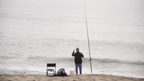 fisherman standing on the sandy shore with the fishing rod, fishing on the ocean with waves. - wide shot