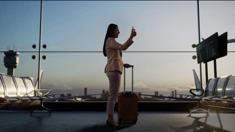 full body back view of asian businesswoman with rolling suitcase in boarding lounge at the airport, waving hand having video call on smartphone, airplane takes off outside the window