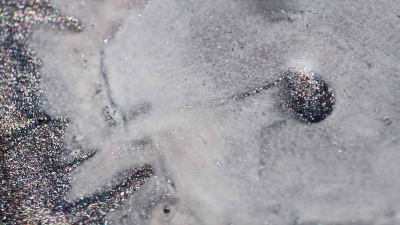 Macro view of glittery grey inks blending as a milky liquid swirls in, creating fluid abstract patterns