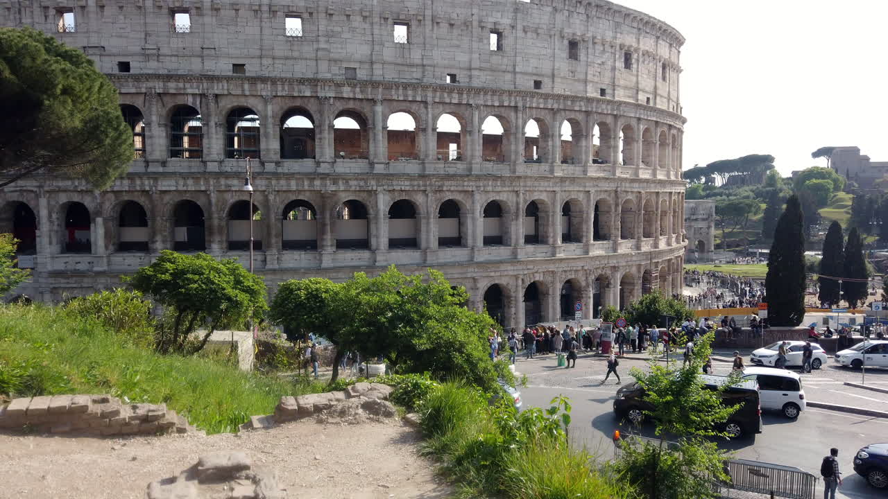 The colosseum from a different side than usual on a sunny spring day. There are few cars parked on the sides of the street. Cars are passing by. People are walking on the sidewalk.