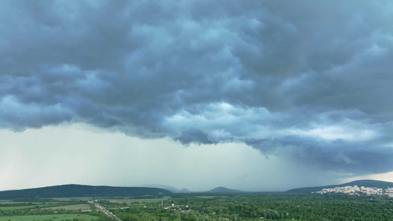 An aerial drone video captures a dramatic storm brewing over a vast landscape. Dark clouds loom ominously, suggesting severe weather, while rolling hills and distant towns dot the green fields below.