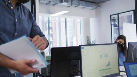 Diverse-male-and-female-colleague-wearing-face-masks-elbow-bumping-at-desk-in-busy-office