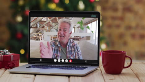 hombre caucásico de alto nivel saludando en una videollamada en una computadora portátil, con decoraciones de navidad y árbol