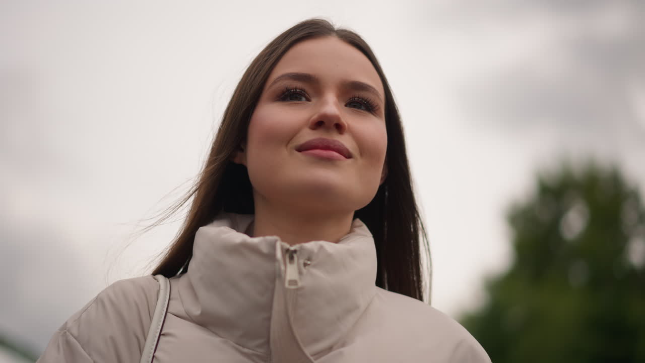 caucasian woman outdoors wearing beige jacket, poses as model, walks as commuter, reflects as student, smiles as friend, gazes toward cloudy sky, wind in long hair, park trees blurred background, soft