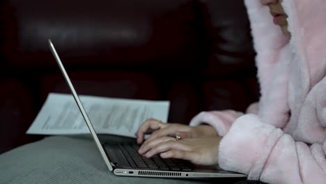 Persona-Con-Bata-Rosa-Escribiendo-En-Una-Computadora-Portátil,-Sentada-En-El-Interior,-Concepto-De-Trabajo-Desde-Casa,-Poca-Luz,-Ambiente-Acogedor