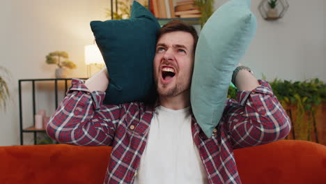 irritated angry man covers ears with cushions annoyed by noisy loud music neighbors repair work