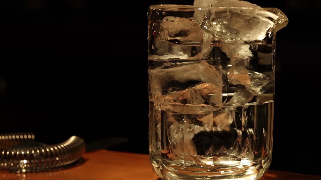 Close-up of hands stirring ice in a glass with a strainer nearby on a wooden surface.