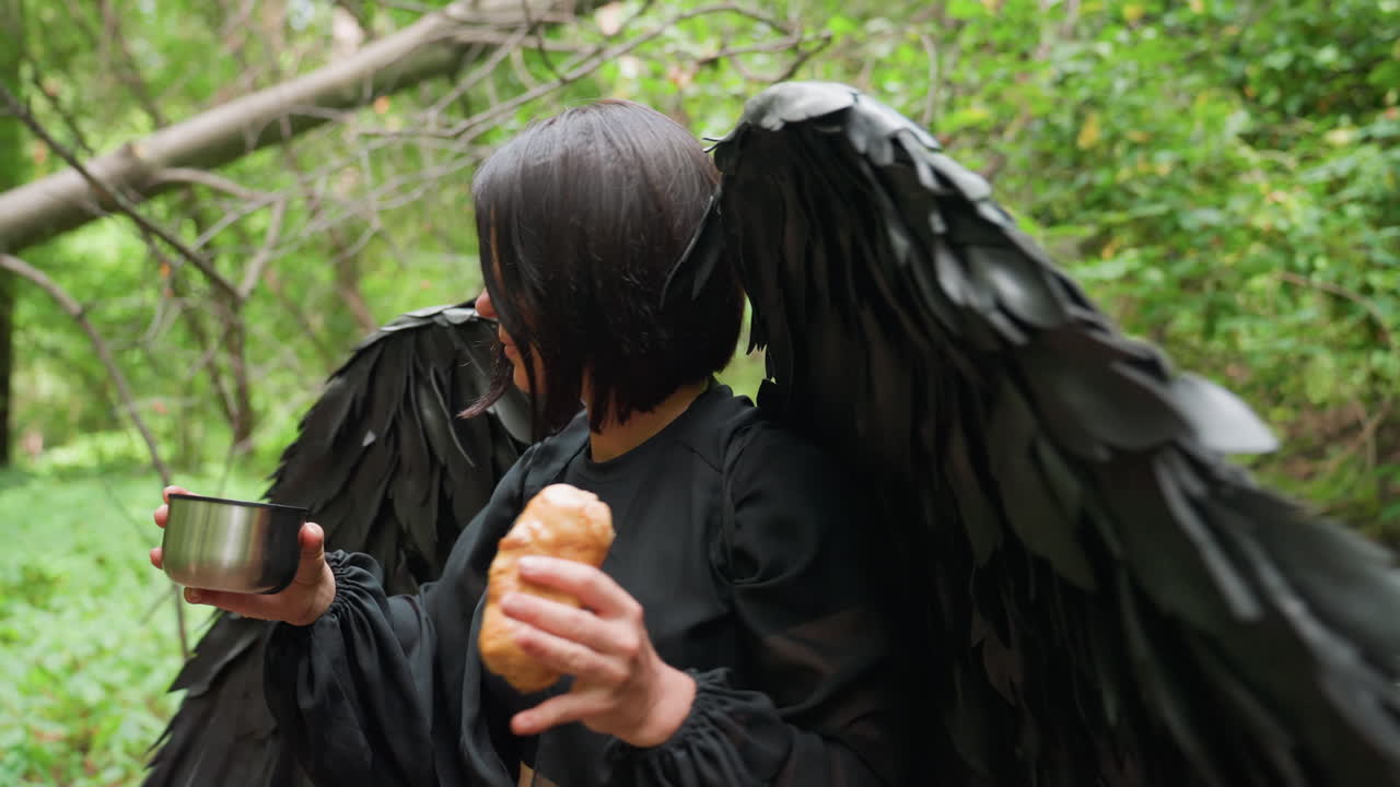 Mysterious black fairy with dark wings gently picks snack and cup from fallen tree in lush green forest, surrounded by sunlight and branches, before walking away with graceful