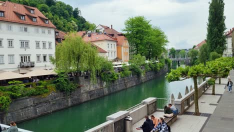 view from one side of the canal to historical buildings on the opposite side with people walking, sitting end enjoying the riverbank and bridge on a cloudy spring day in ljubljana slovenia