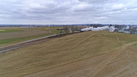 an aerial view of an approaching steam train traveling thru the countryside blowing smoke and steam on a winter day