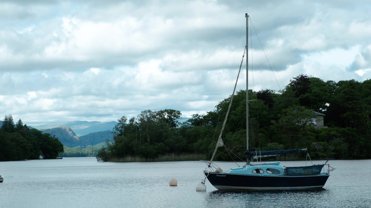 Inquadratura statica di una barca a vela ormeggiata sul Coniston Water nel Lake District, incorniciata da nuvole drammatiche, acqua calma, riva boscosa e rilievi distanti