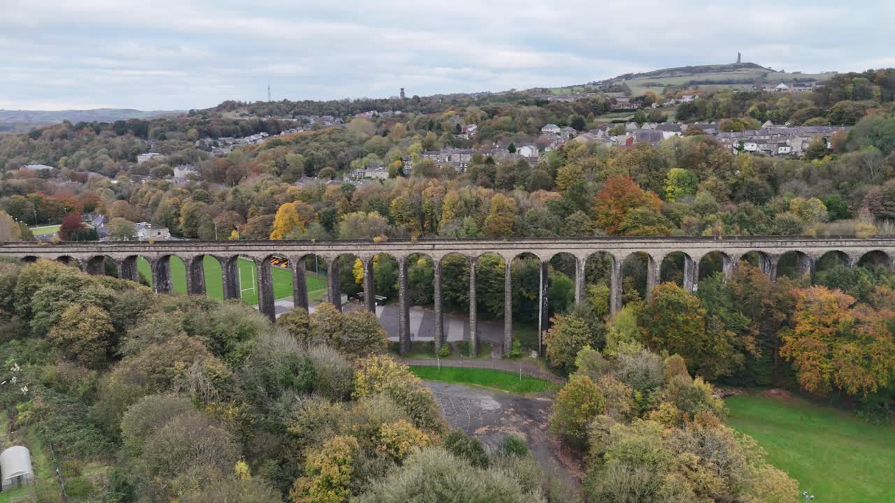 Drone view of Lockwood Viaduct spanning across lush green valley with trees in autumn colors and residential houses in Huddersfield, West Yorkshire, on calm cloudy day