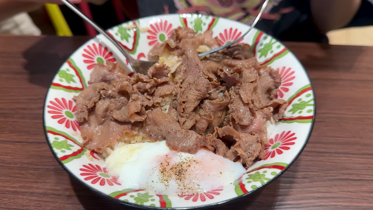 A person uses a spoon and fork to add pickled ginger onto a beef rice bowl with a soft-boiled egg, under bright indoor lighting at a wooden table