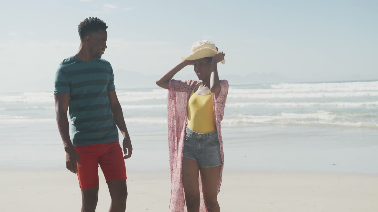 Happy african american couple walking with hat on sunny beach