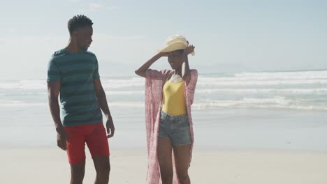 feliz pareja afroamericana caminando con sombrero en una playa soleada
