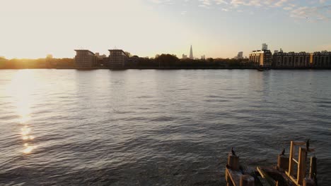 Aerial-flyover-River-Thames-during-sunset-with-water-reflection,-cruising-boat-and-skyline-silhouette